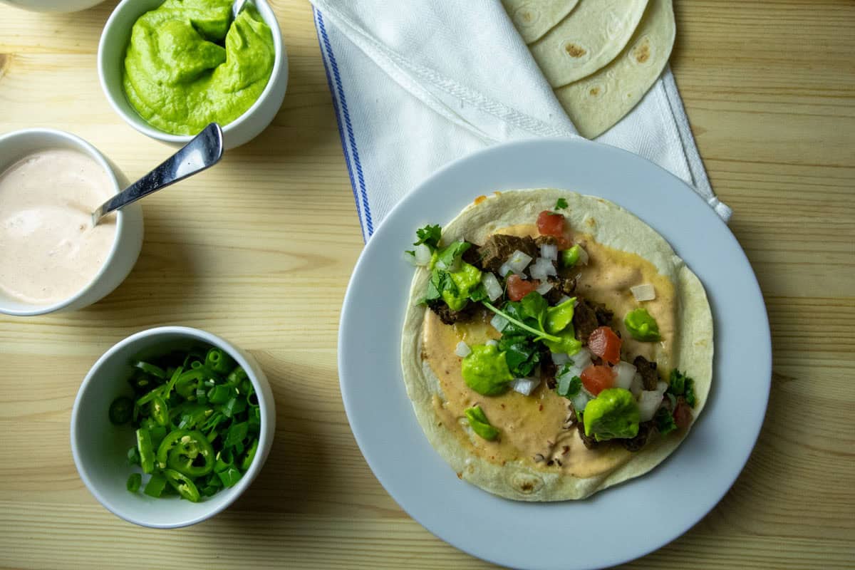 Assembled vampiro taco on a plate with tortillas, sliced serrano peppers, and tomatillo avocado sauce in the background. 