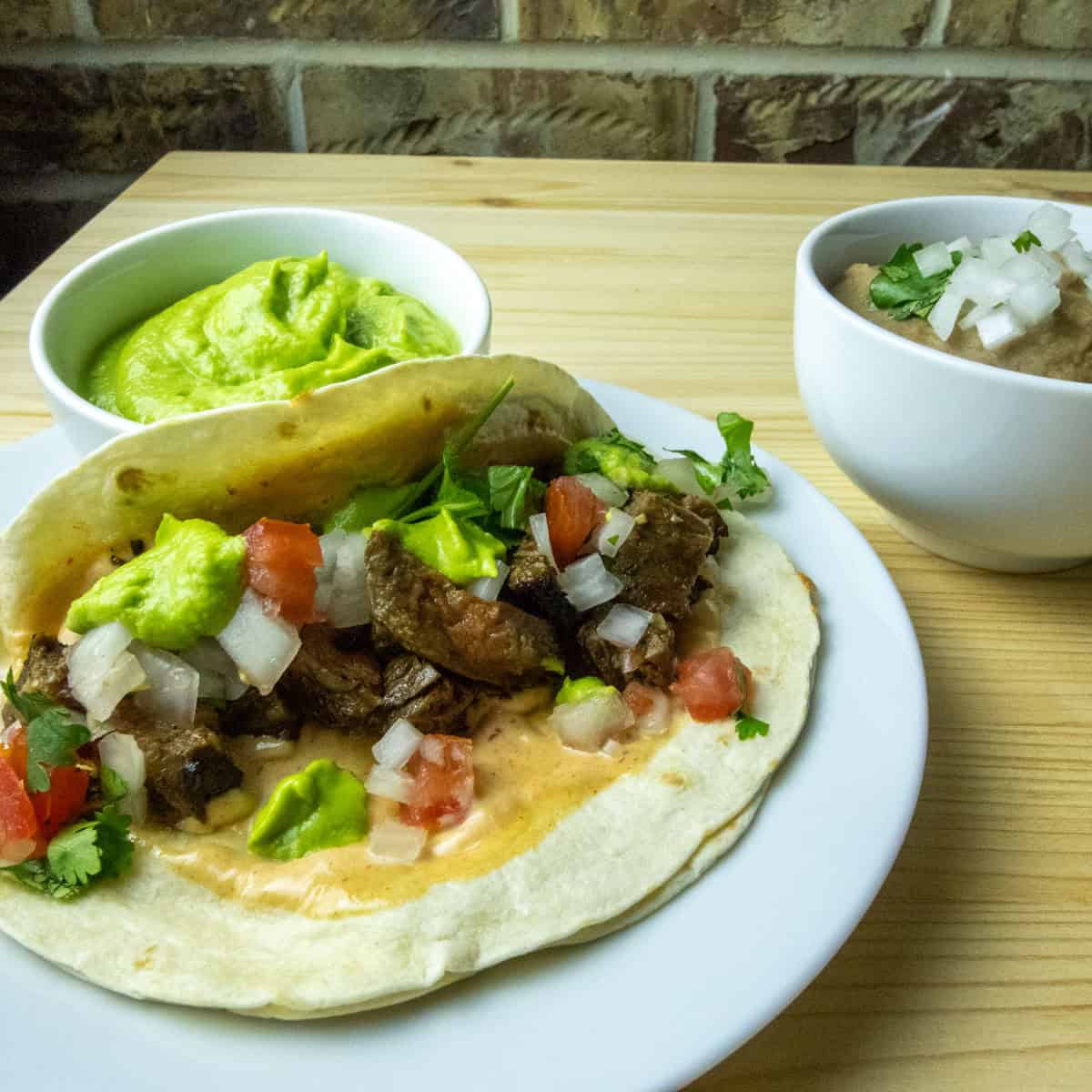 Vampiro taco on a plate with pico de gallo and chipotle mayo with tomatillo avocado salda and refried beans in the background.