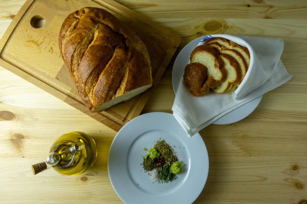 Herbs on a plate with a bottle of olive oil beside it with a loaf of bread and slices in the background.
