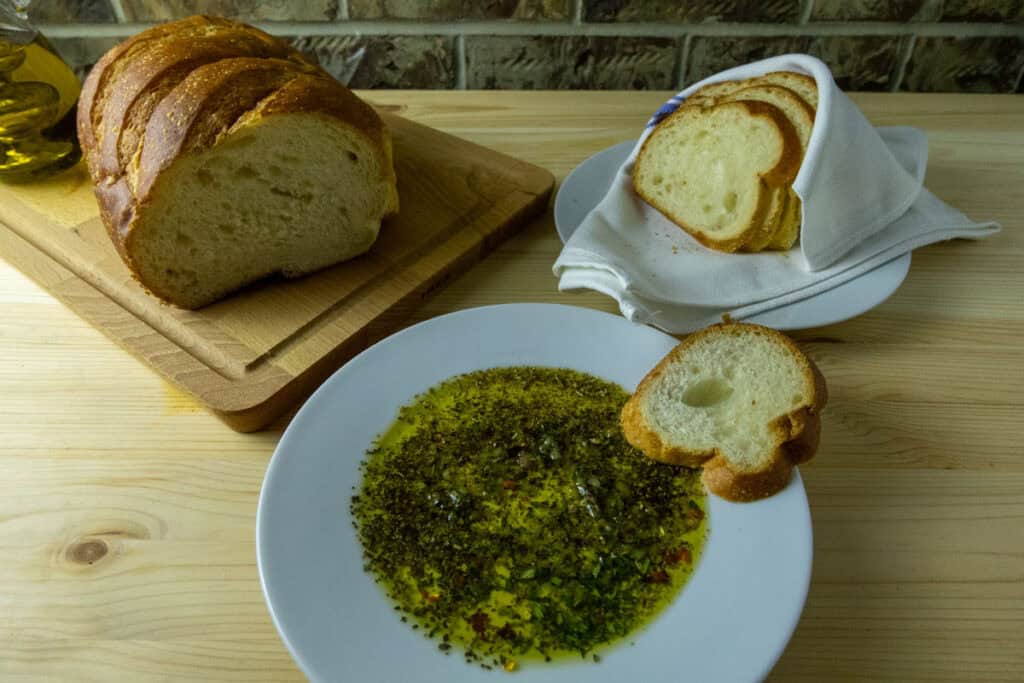 Olive oil herb dip on a plate with a slice of bread on the side with a loaf and slices in the background.
