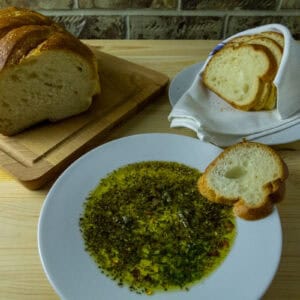 Olive oil herb dip on a plate with a slice of bread on the side with a loaf and slices in the background.
