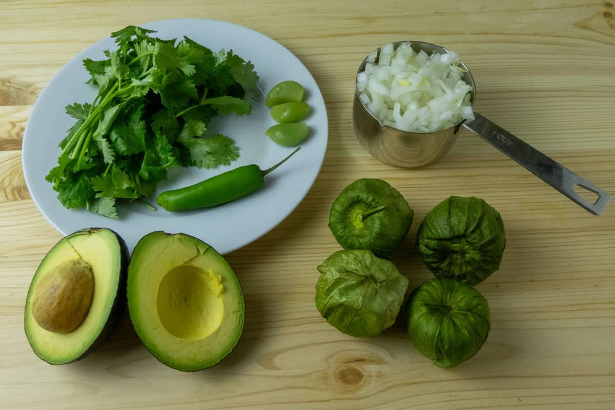All ingredients to make tomatillo salsa including cilantro, Serrano pepper, tomatillos, avocado, and white onion,