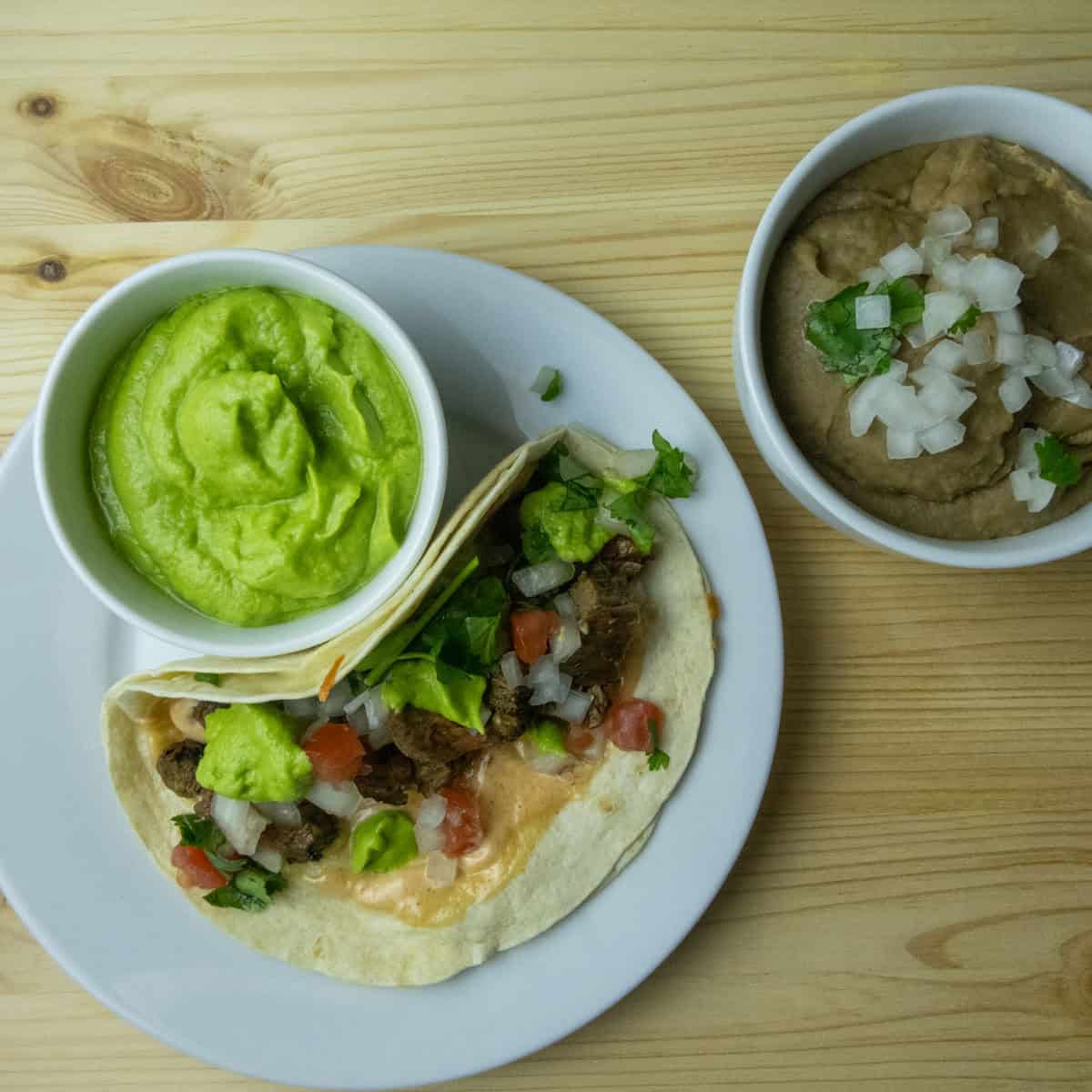 White plate with a Vampiro Taco and a bowl of Tomatillo Avocado Salsa and a small bowl of refried beans.