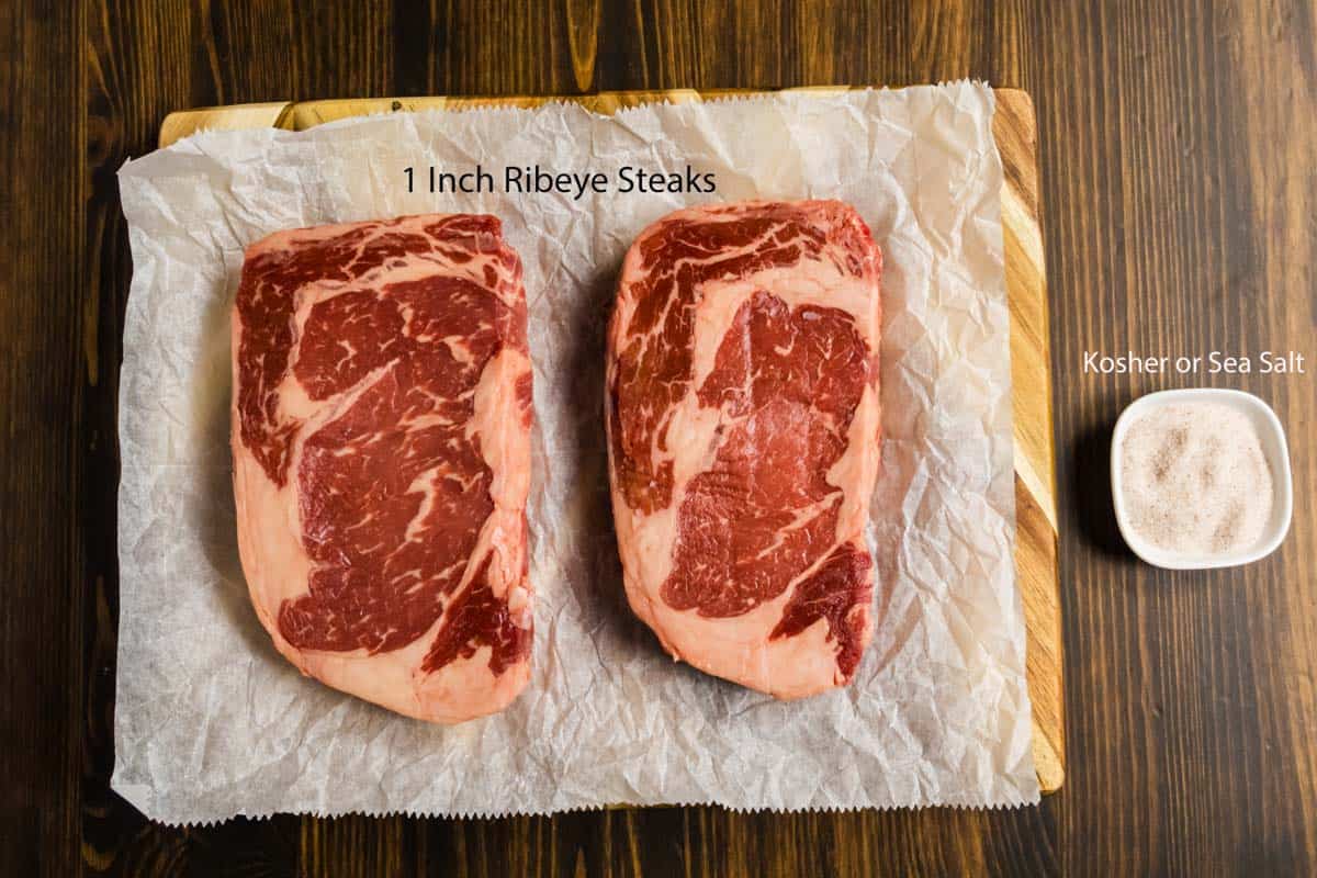 Two ribeye steaks on parchment paper with salt in a container to the side.