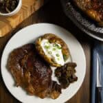 Dry brined steak on a plate with a loaded baked potato and mushrooms and onions.