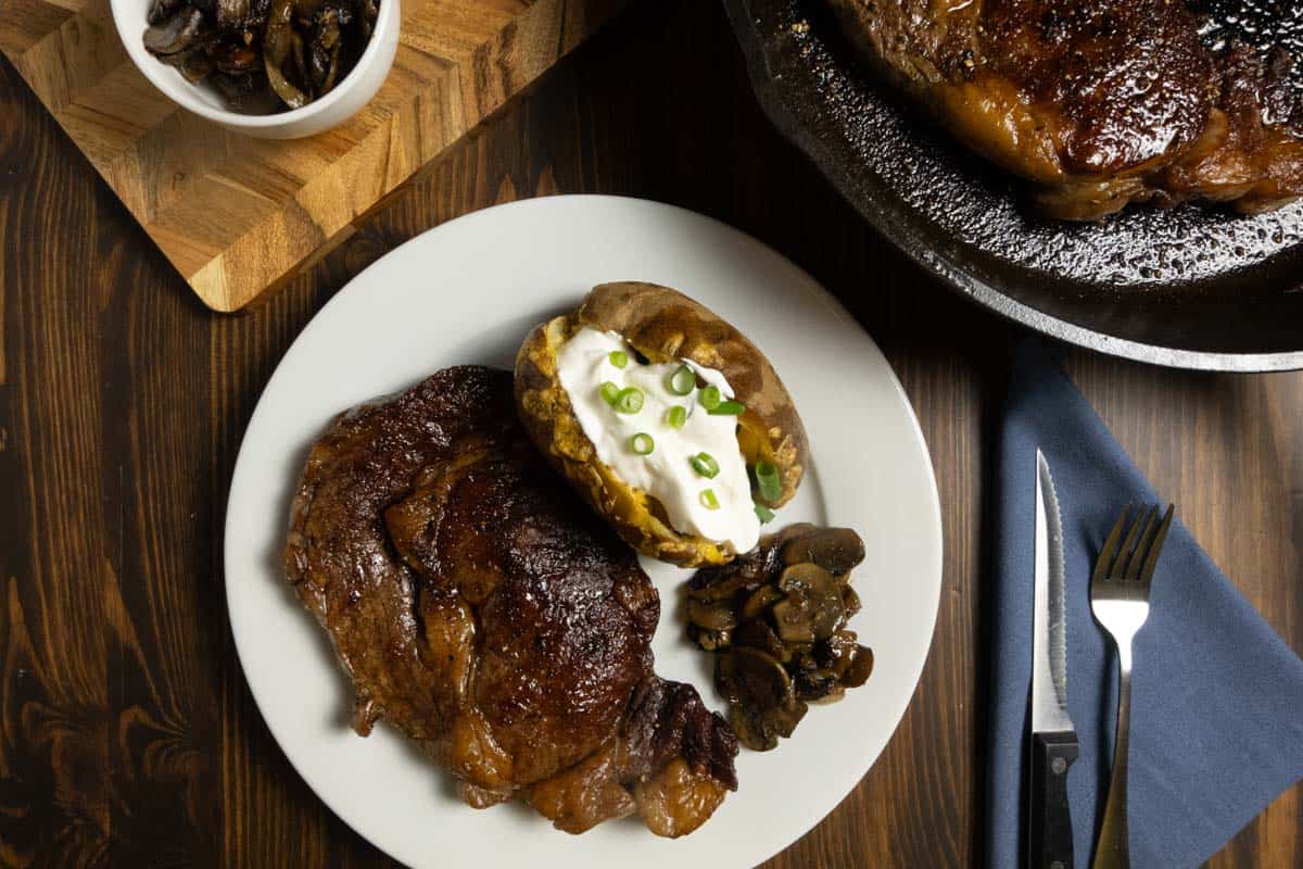 Dry brined steak on a plate with a loaded baked potato and mushrooms and onions.