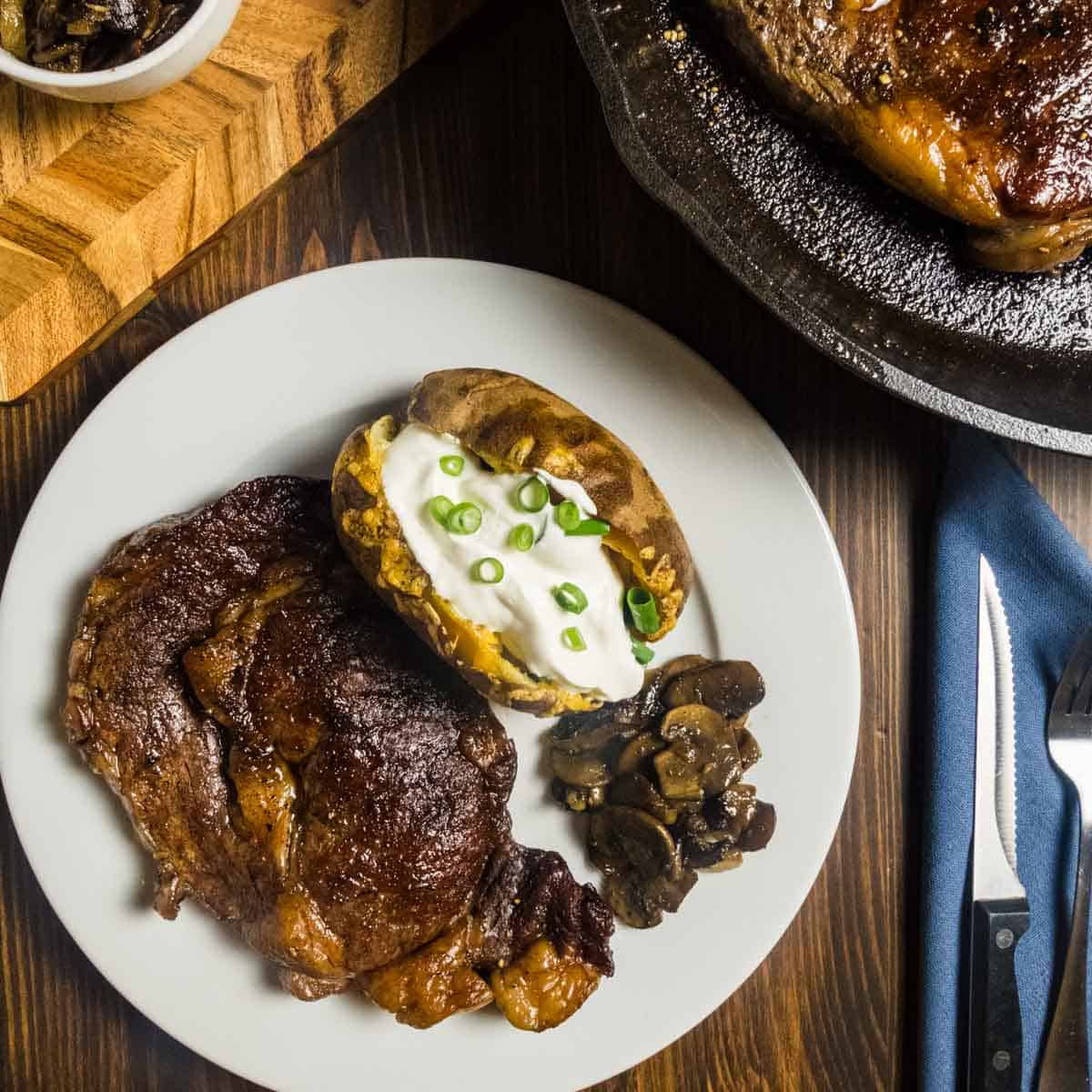 Dry brined steak on a plate with mushrooms and onions and a loaded baked potato,