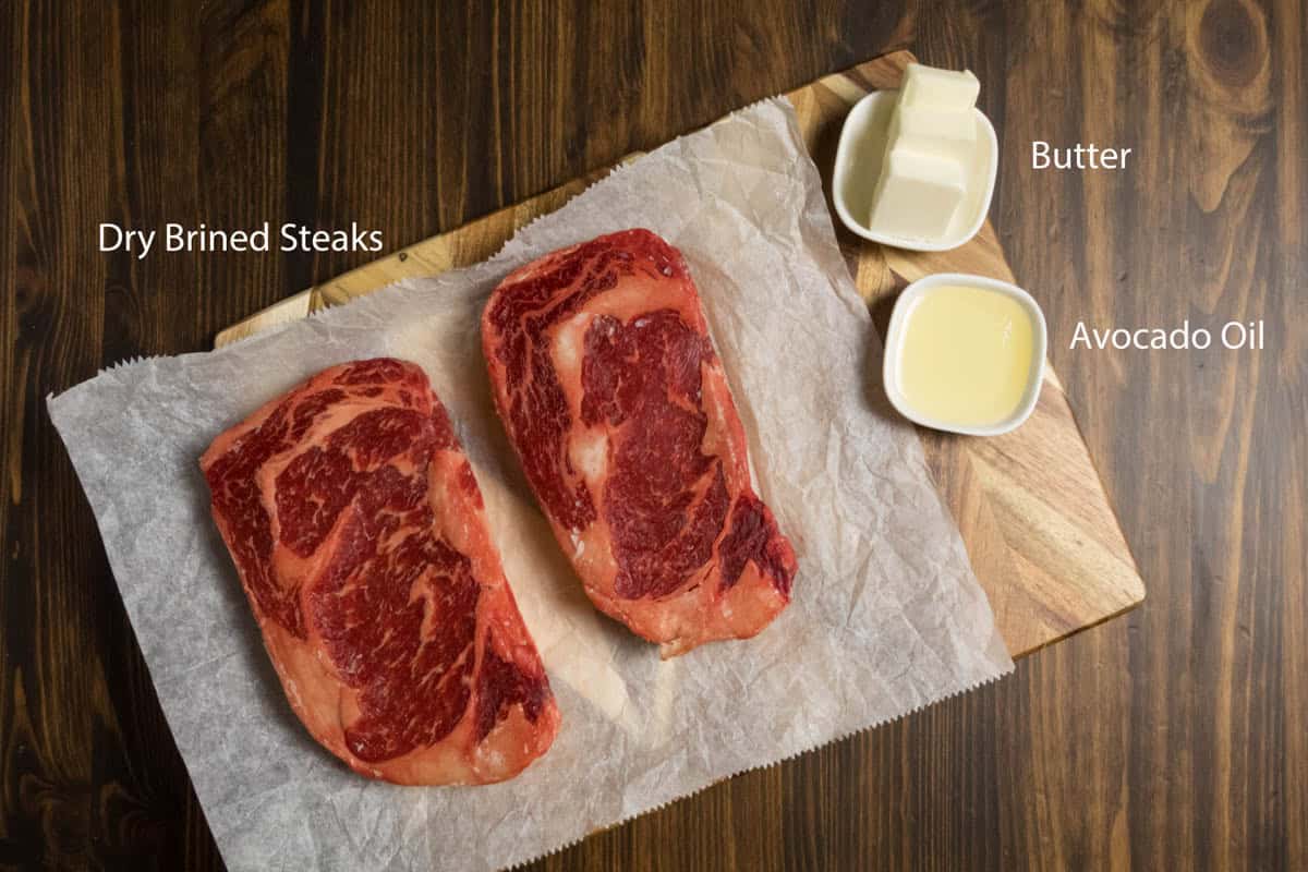 Two dry brined steaks on parchment paper with butter and oil for cooking.
