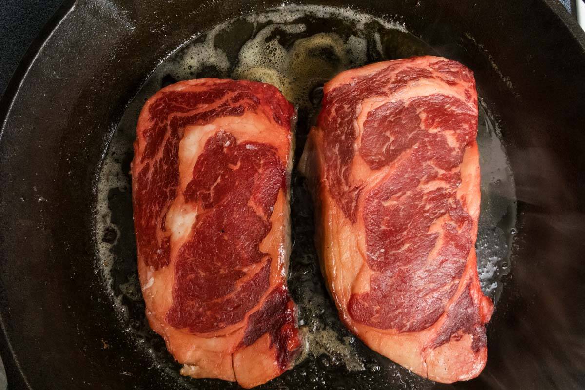 Two steaks cooking in a cast iron skillet.