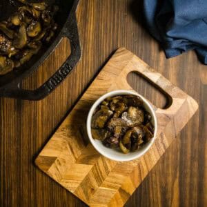 A bowl of caramelized onions and mushrooms on a cutting board with a cast iron skillet of mushrooms in the back.