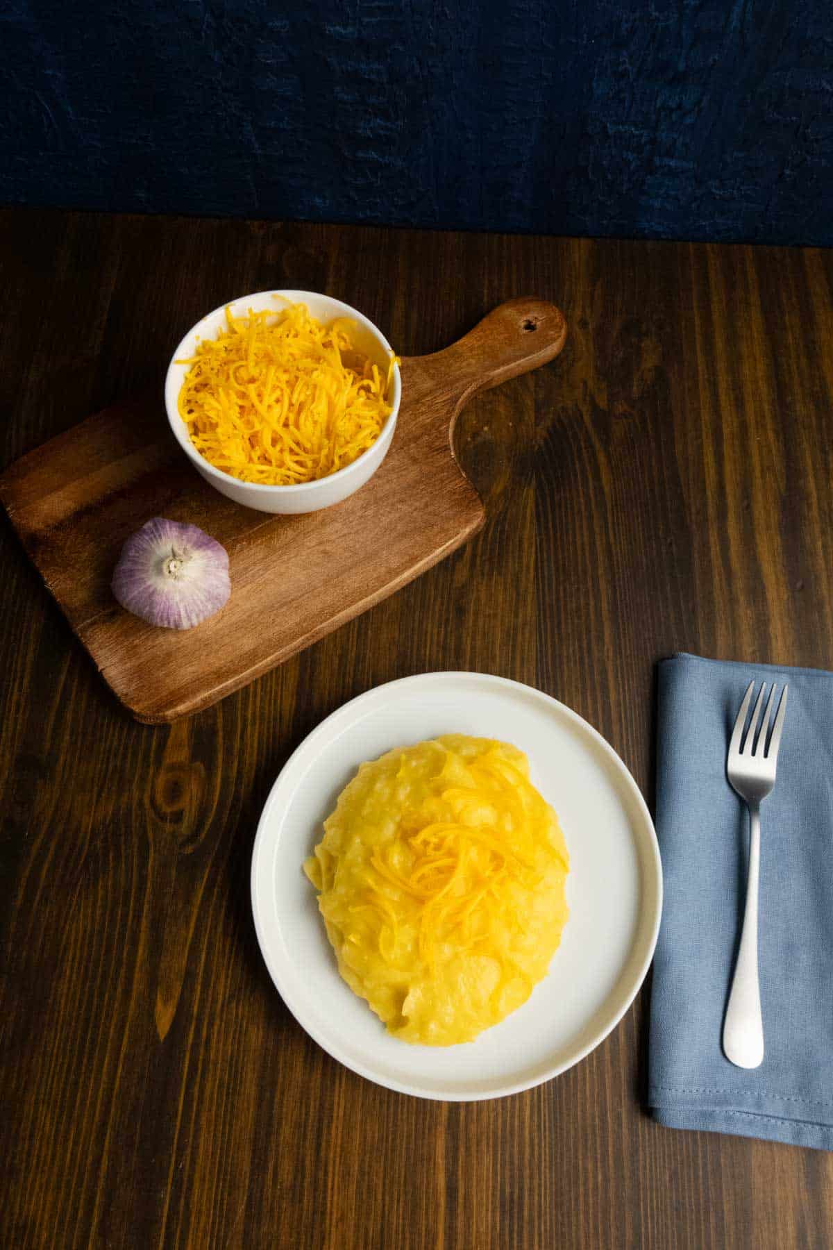A plate with cheesy garlic mashed potatoes with a garlic bulb and cheese in the back on a cutting board.