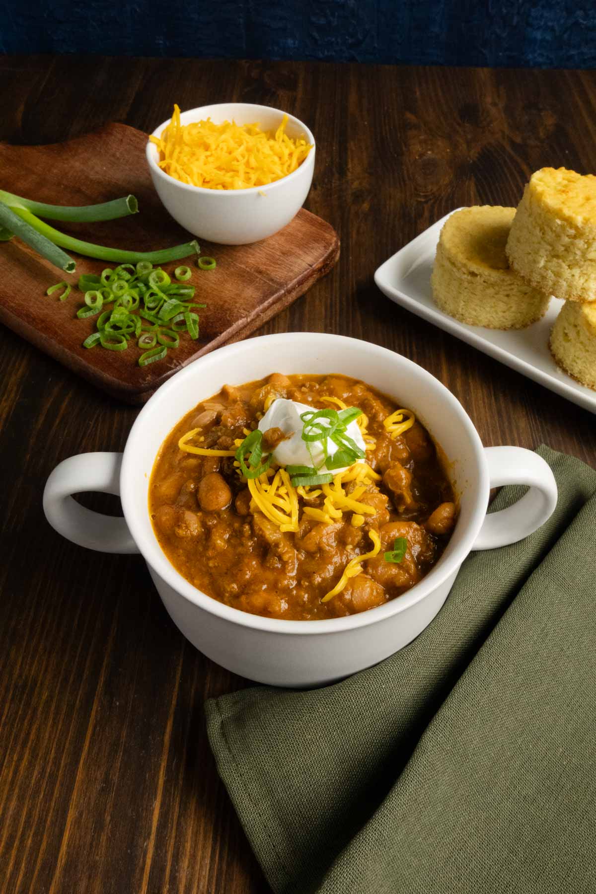 Bowl of chili with cheese, green onions, and cornbread in the background.