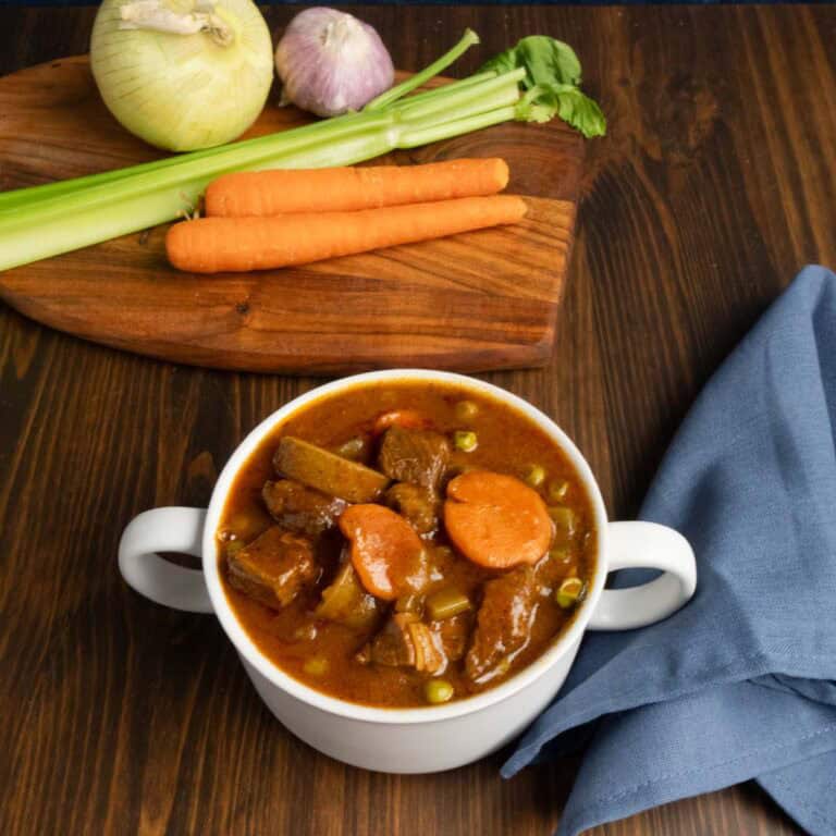 Bowl of stovetop beef stew with onion, carrots, celery, and garlic in the background.