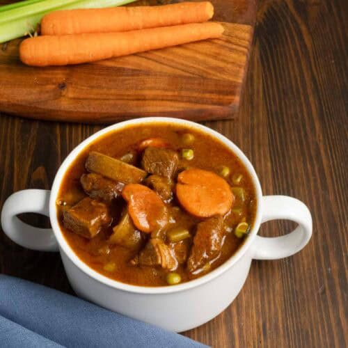 Bowl of beef stew on the stovetop with a blue napkin and carrots and garlic in the background.