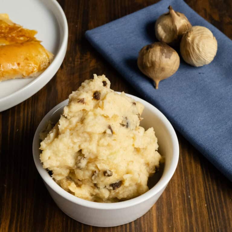 Black garlic mashed potatoes in a bowl with turkey and black garlic bulbs in the background.
