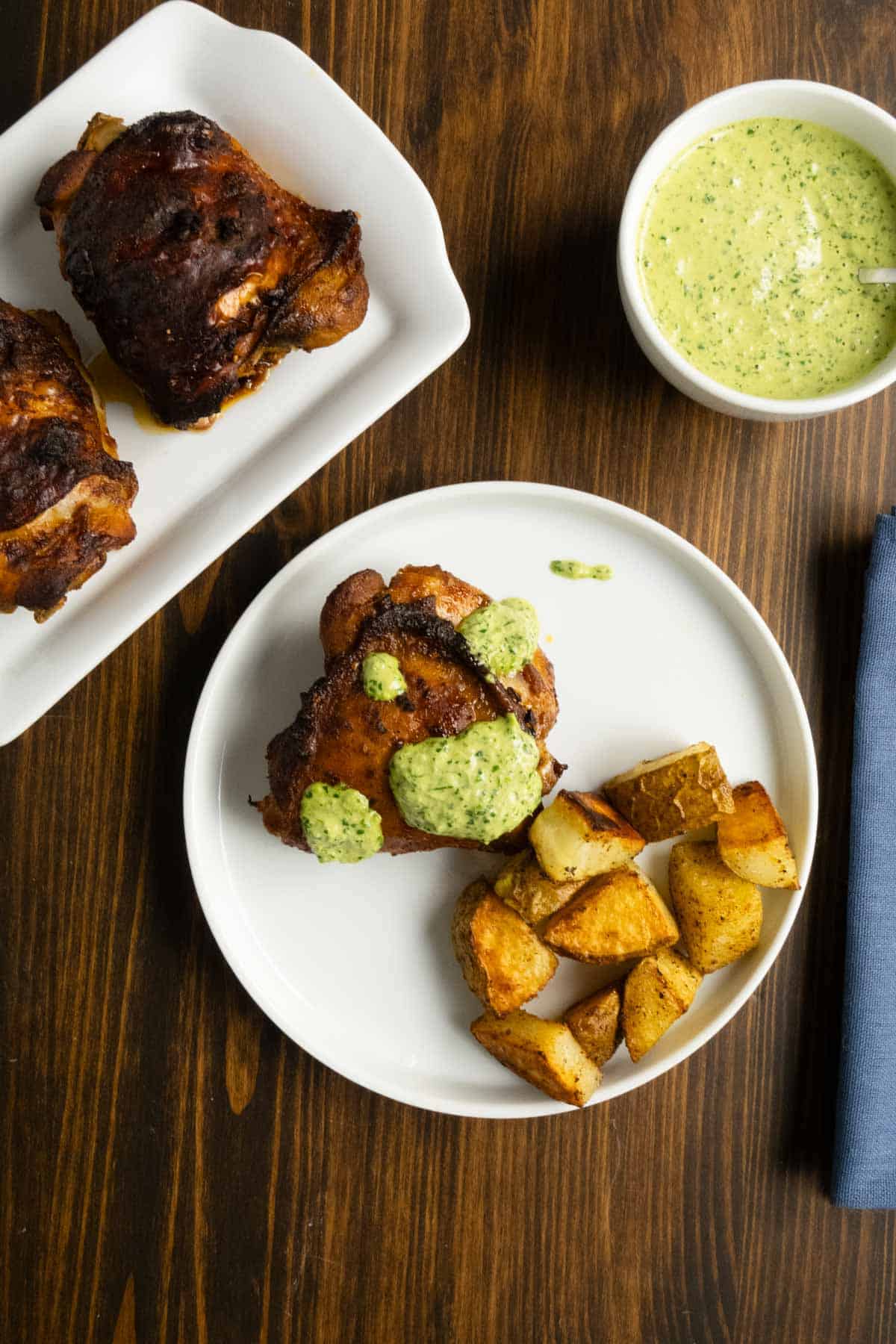 A plate of Peruvian chicken with green sauce and extra crispy roasted potatoes with extra chicken and sauce in the background.