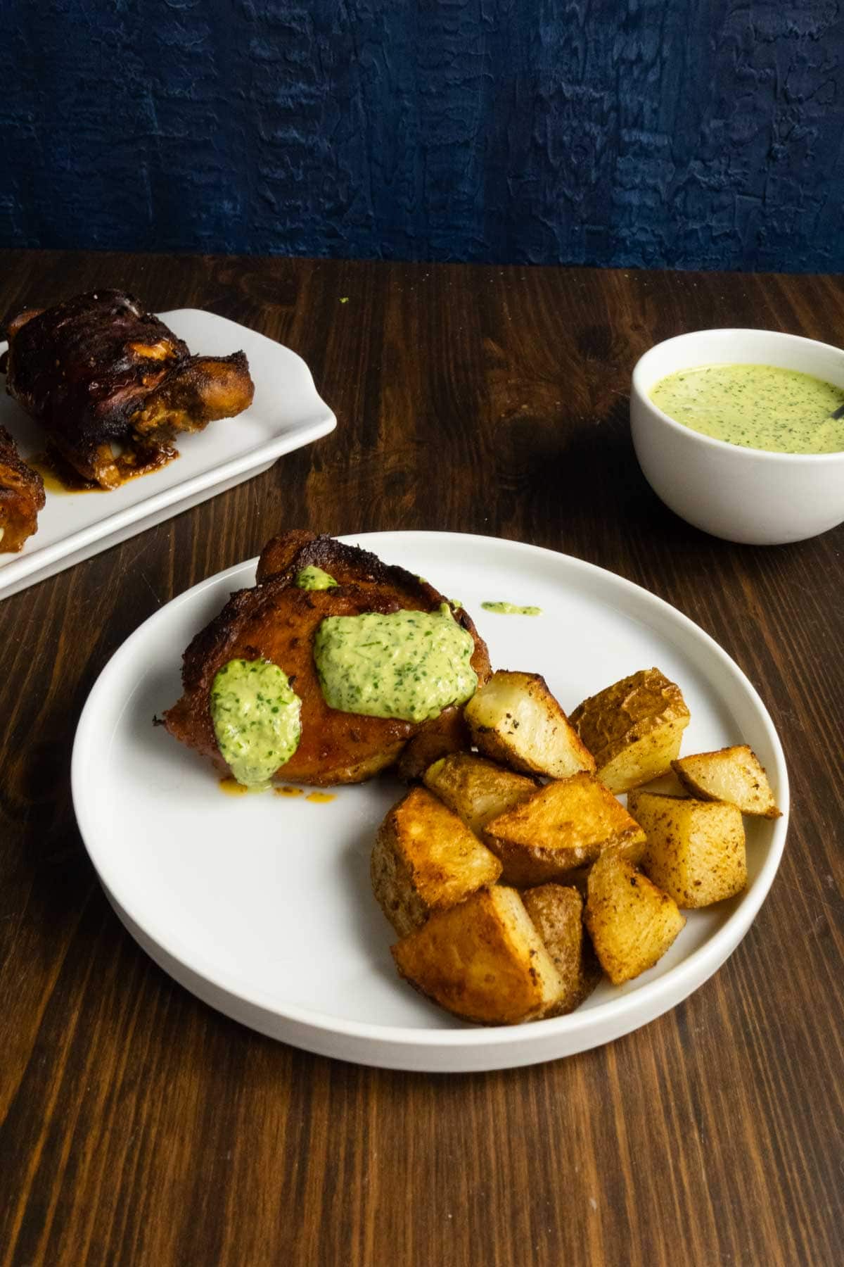 Green sauce is a bowl with a plate of chicken thighs and roasted potatoes.