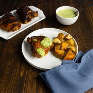 Plate of cooked chicken thighs with green sauce and roasted potatoes.
