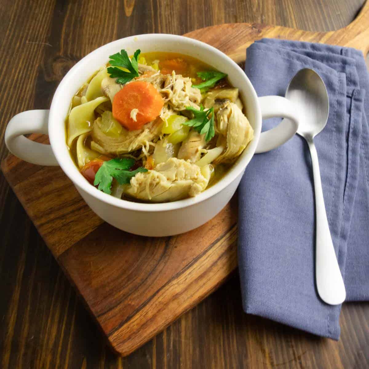 A bowl of Grandma's chicken noodle soup on a cutting board with a blue napkin and a spoon.