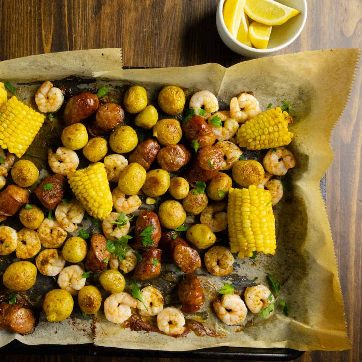 A sheet pan with shrimp, sausage, potatoes, and corn with lemon wedges.
