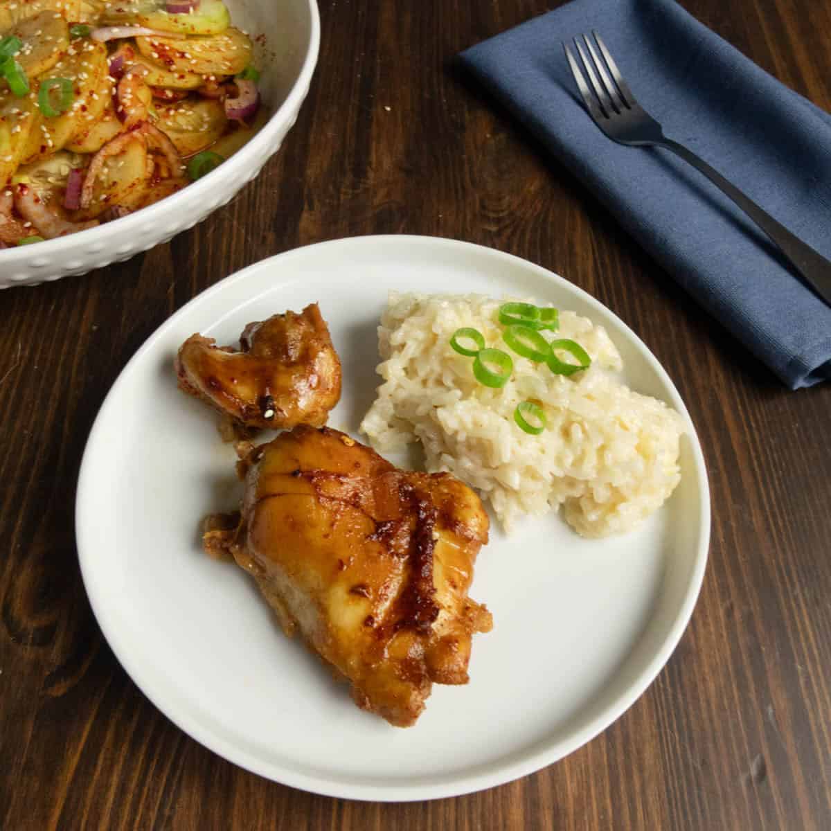 A plate with Spicy Asian Boneless Skinless Chicken Thighs and ginger coconut rice with a Korean cucumber salad in the back and a fork on a blue napkin.