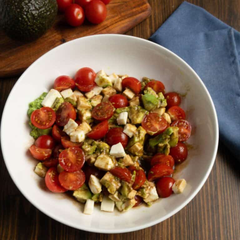 A bowl with tomato, avocado, mozzarella salad with an avocado and whole cherry tomatoes on a cutting board.