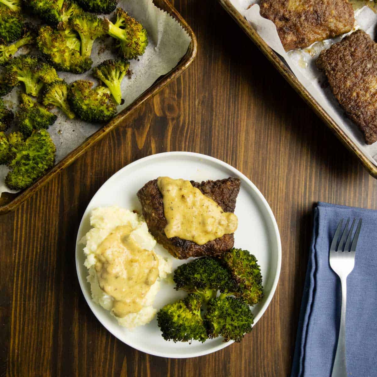 Roasted broccoli on a plate with cube steak, mashed potatoes, and gravy with broccoli in the background.