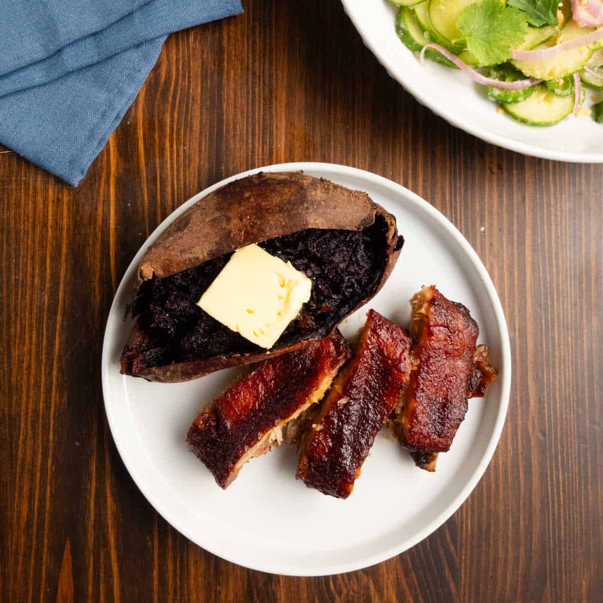 A baked purple sweet potato on a white plate with chipotle barbecue glazed dry brined pork ribs.