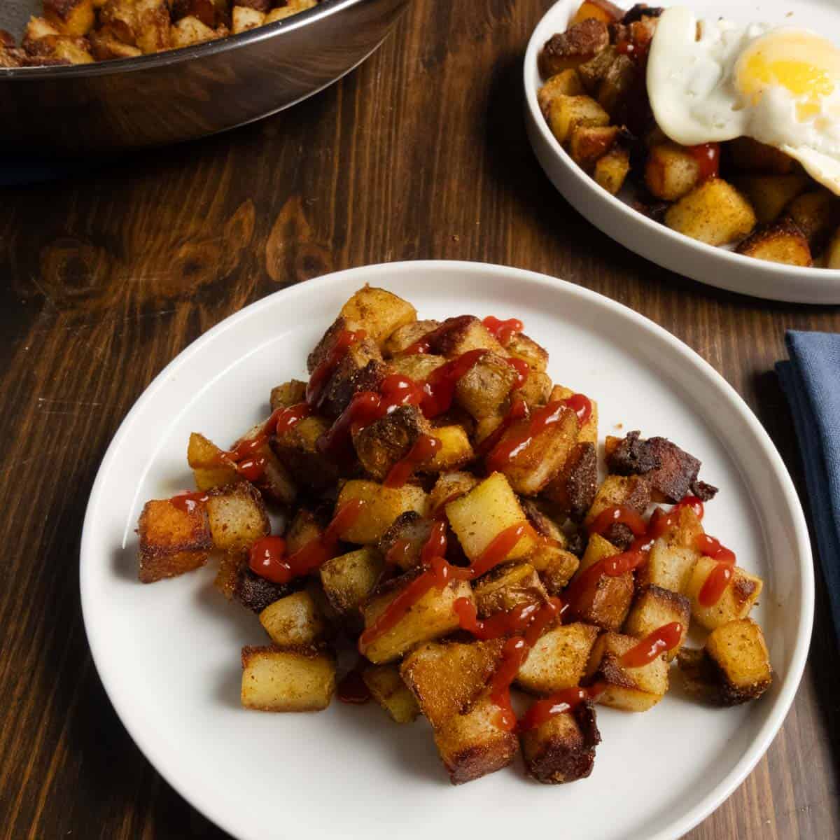 Crispy home fries on a plate with ketchup on it with a fried egg in the background.