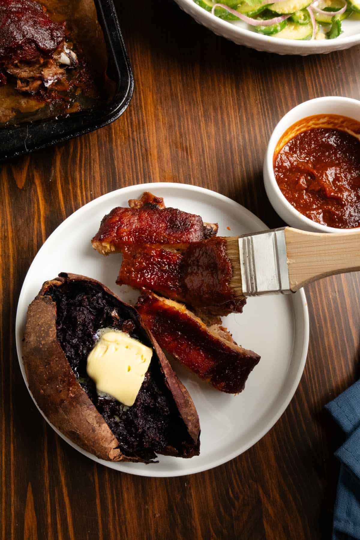 Plate with ribs getting an extra brushing of barbecue sauce with a purple sweet potato and Thai cucumber salad in the background.