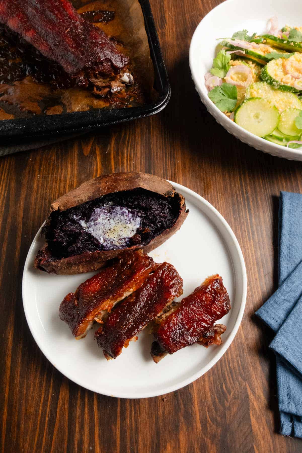 Dry brined pork ribs with a homemade chipotle barbecue sauce, a baked purple sweet potato, Thai cucumber salad, and a pan with ribs on it.