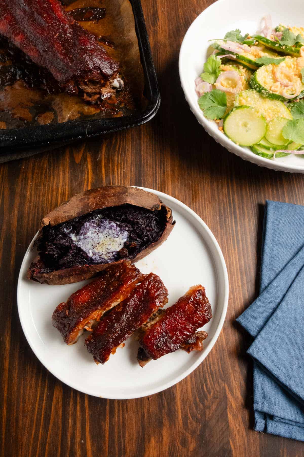 Finished ribs on a plate with Thai cucumber salad, a tray of ribs, and a purple sweet potato.