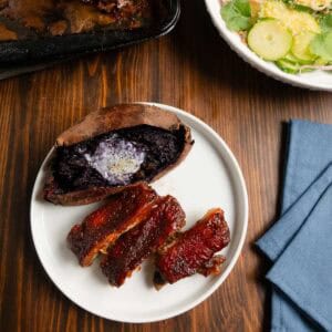 Dry brined pork ribs cooked in the oven on a plate with chipotle barbecue sauce and a purple sweet potato.