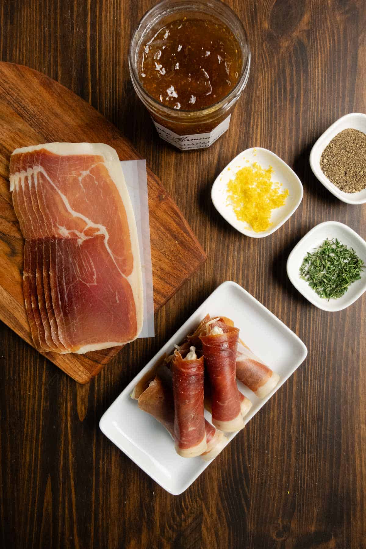 Prosciutto cheese rolls on a white platter with thyme, lemon zest, and fig jam on the side.