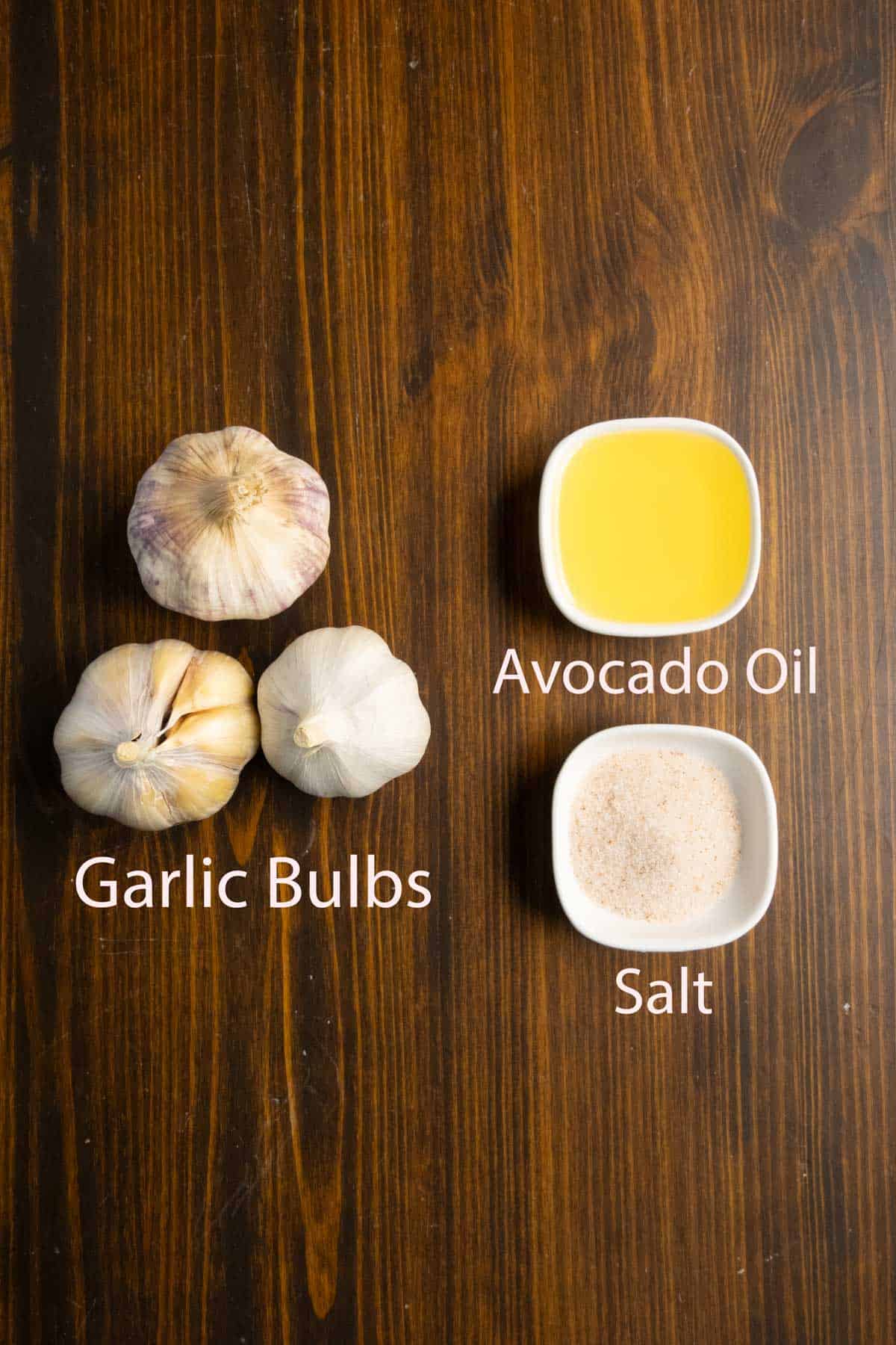 Three garlic bulbs, avocado oil and salt on a table ready to make roasted garlic.
