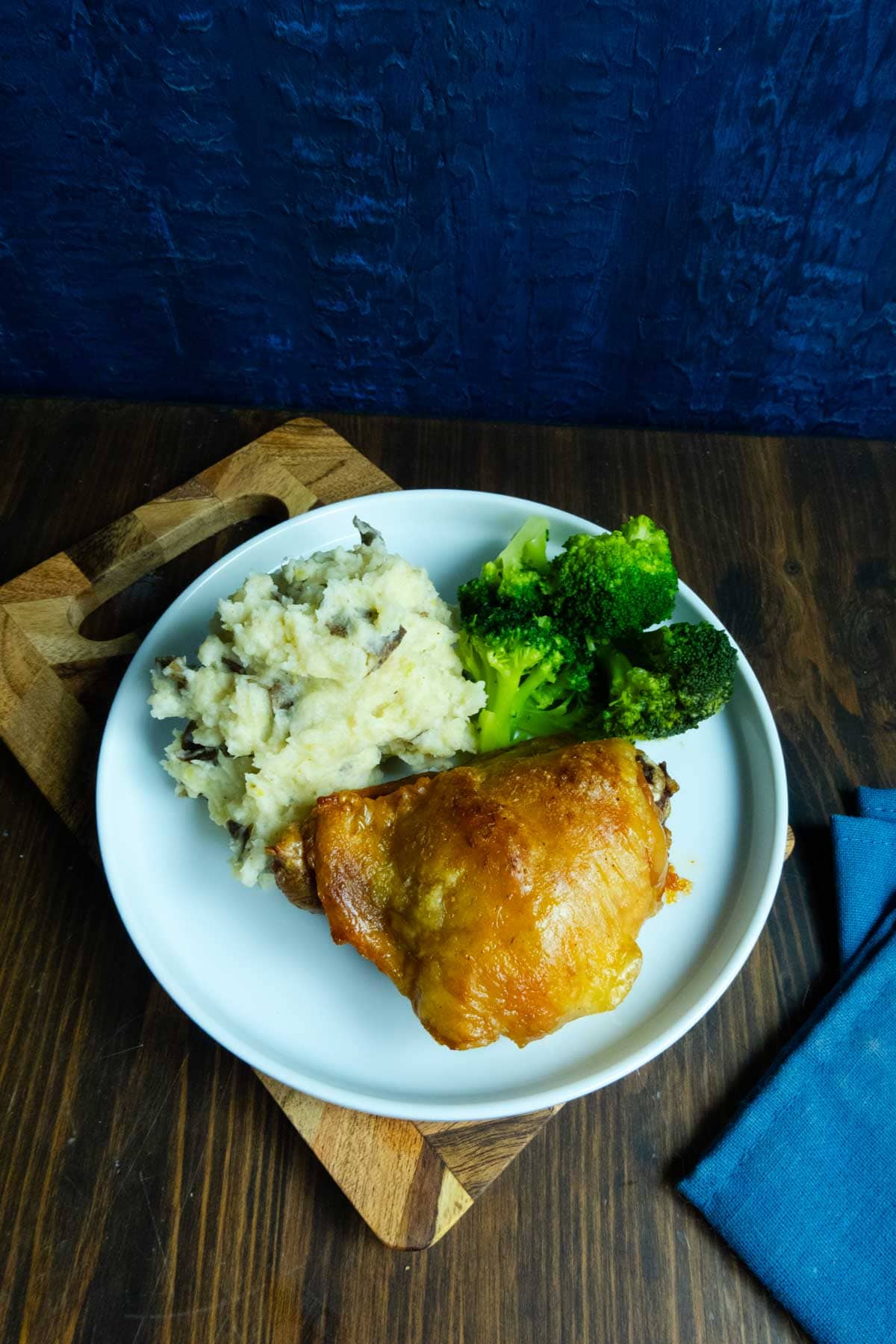 Dry brined chicken thigh on a plate on a cutting board with a blue napkin and mashed potatoes and broccoli.