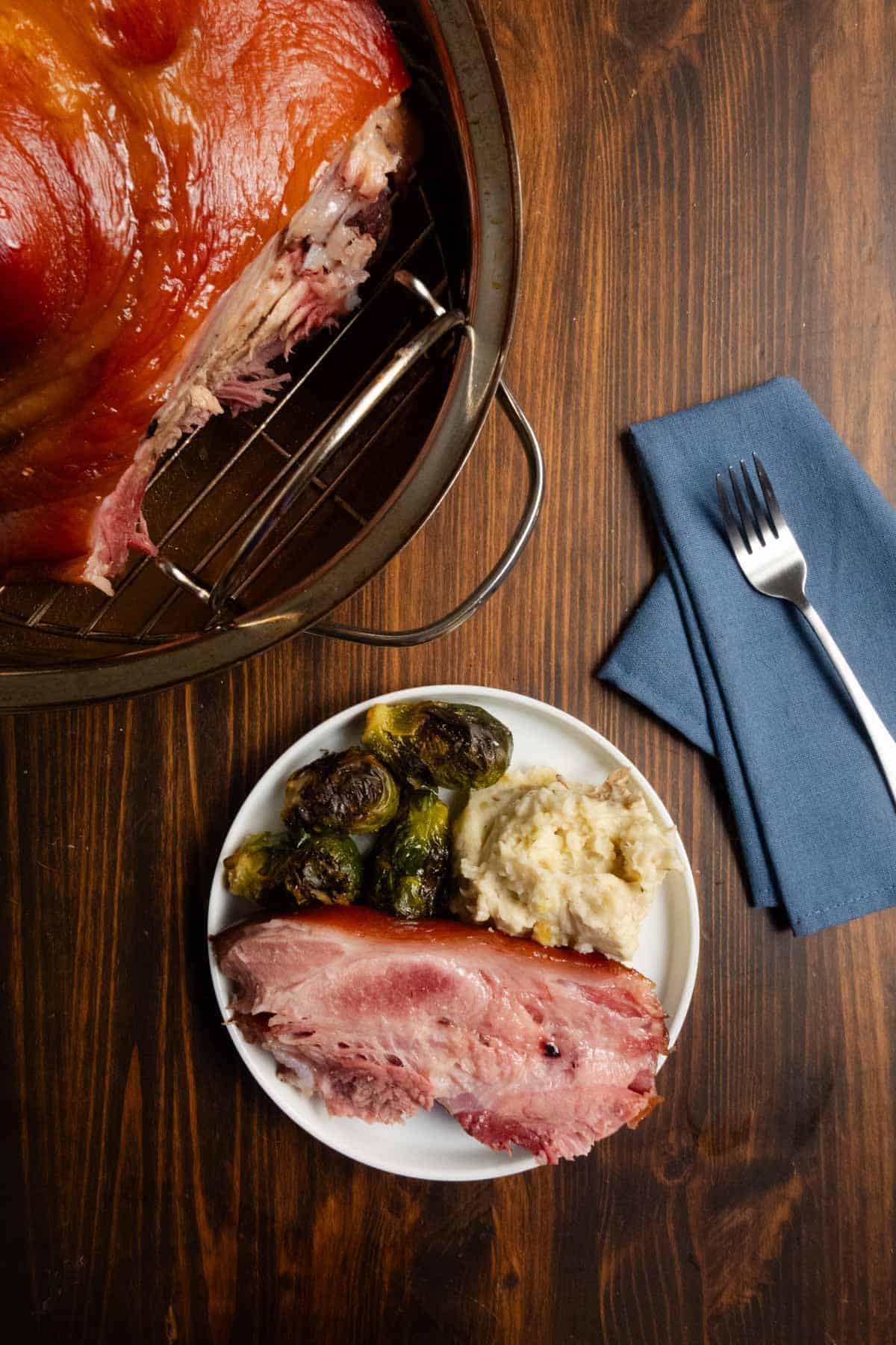 A slice of home cured ham on a plate with mashed potatoes and brussels sprouts with a whole ham in the background.