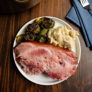 Sliced ham on a plate with brussels sprouts and mashed potatoes.