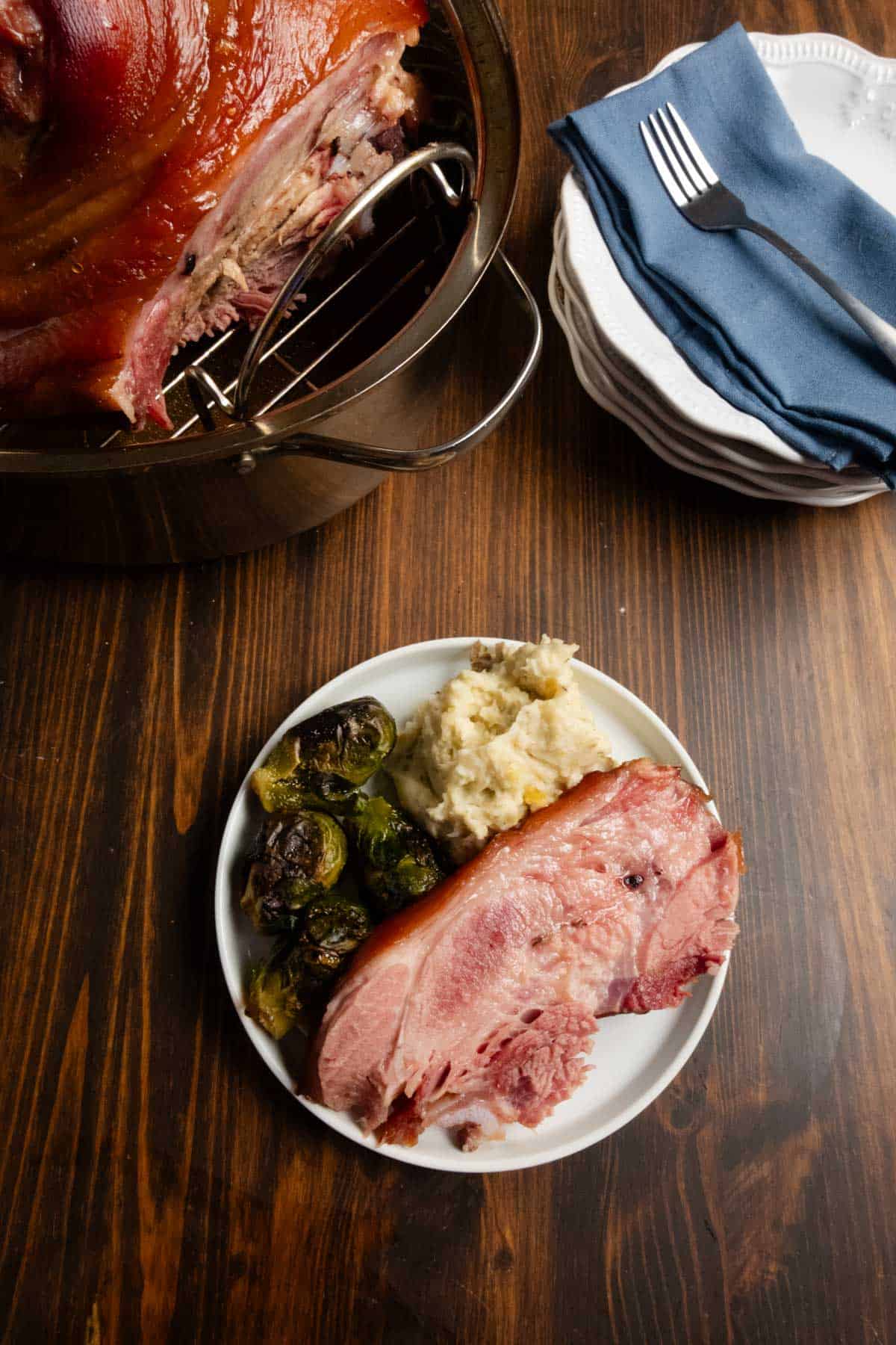 A plate of cured ham with brussels sprouts with a whole ham in the background with plates and napkins.